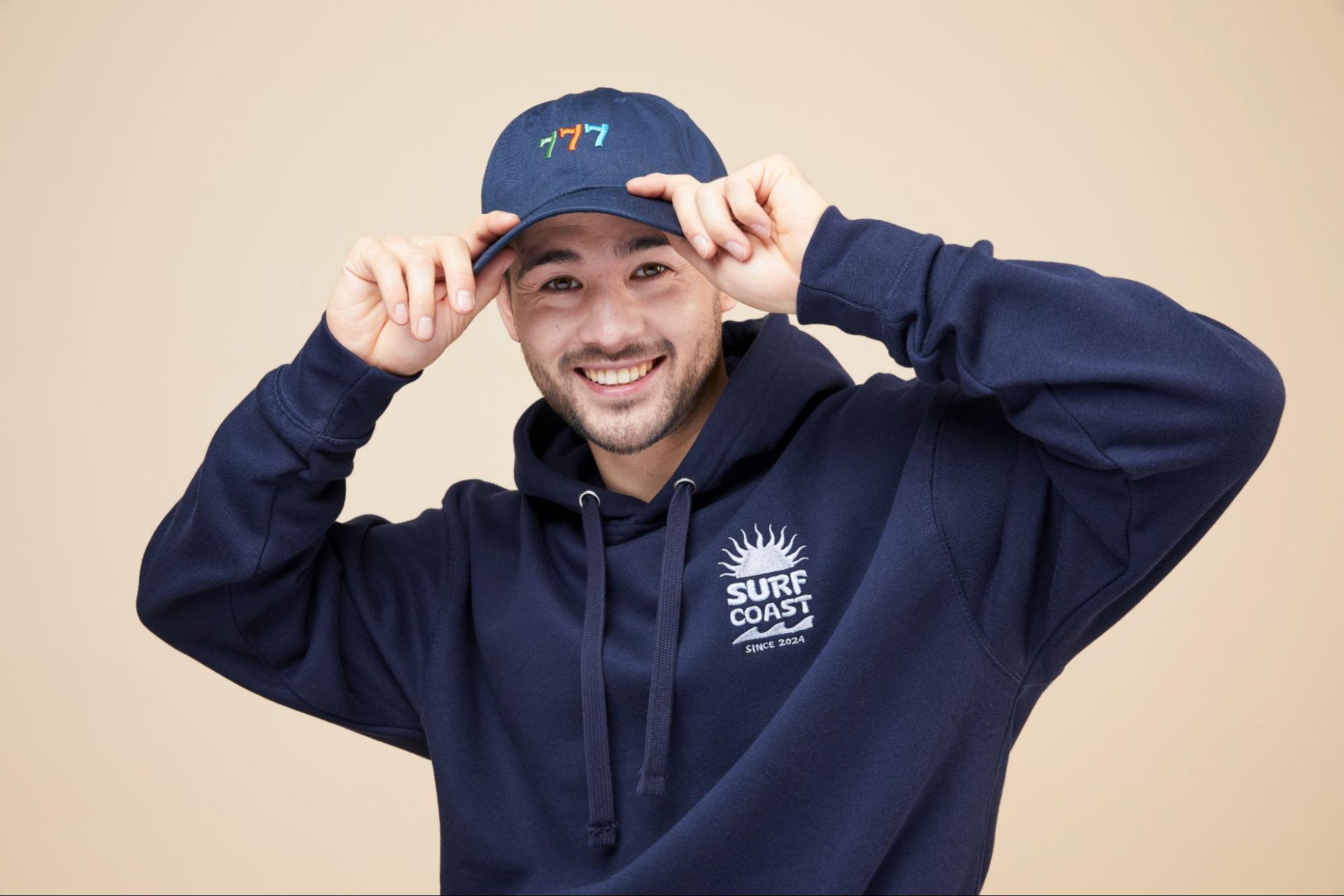 A smiling person wearing a navy embroidered hoodie and cap, showcasing detailed embroidered designs on both garments.